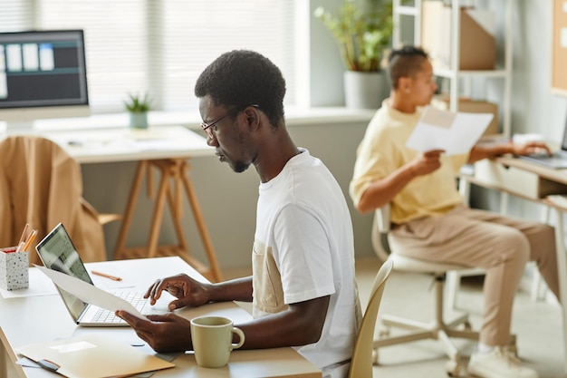 Side view portrait of black young man wearing glasses and using laptop at office workplace copy spac