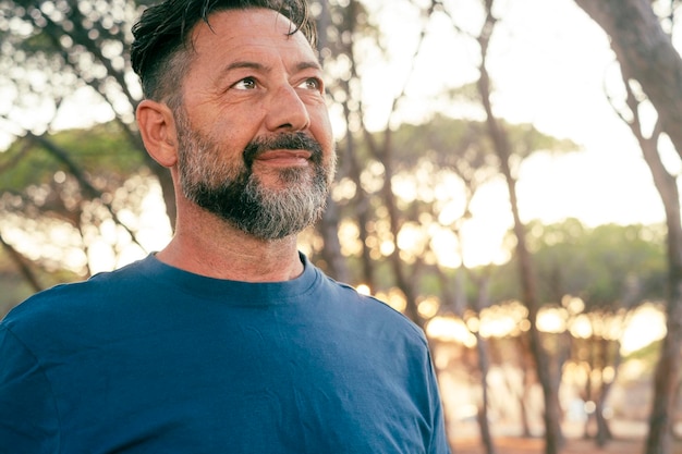 Side view portrait of boomer generation male smiling and looking in front with confident expression and trees forest in background People and outdoor leisure activity alone Casual blue tshirt