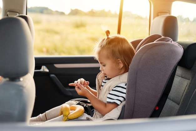 Side view portrait of cute charming baby girl sitting in a car in safety chair playing alone holding bananas wearing striped t shirt traveling