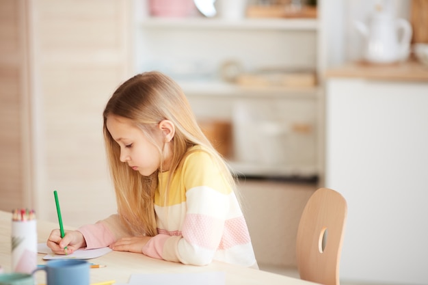 Side view portrait of cute little girl drawing pictures or doing homework while sitting at table in home interior, copy space