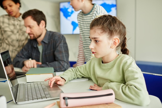 Side view portrait of cute schoolgirl using laptop in modern school classroom with kids in backgroun