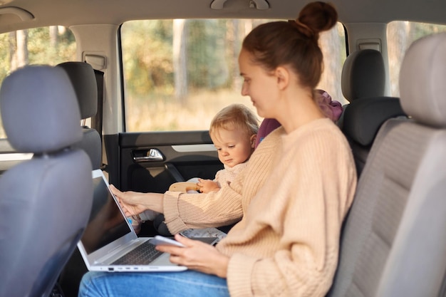 Side view portrait of dark haired woman with bun hairstyle wearing sweater working on laptop while traveling with her daughter with baby seat kid playing while mother doing her work