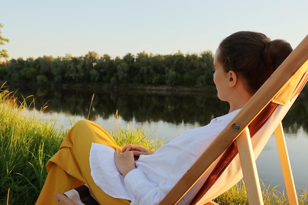 Side view portrait of dark haired young adult woman sitting in the folding chair at the picturesque bank of the river enjoying nature looking away resting alone