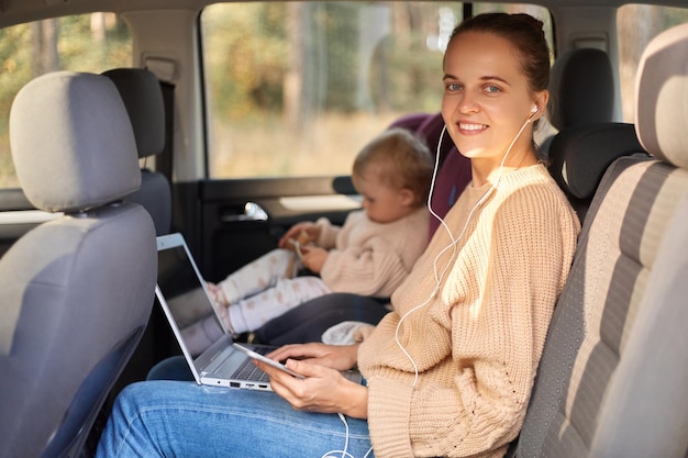 Side view portrait of delighted woman working on notebook while sitting with her baby daughter in safety chair on backseat of auto looking at camera with happy expression listening music