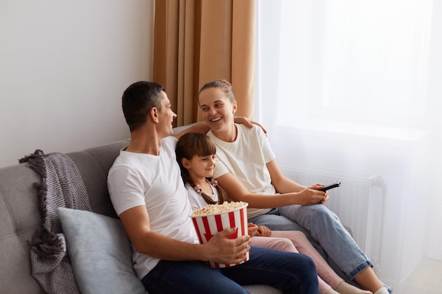 Side view portrait of funny family wearing casual white tshirts sitting on sofa with their daughter and watching movie husband hugging her wife and smiling