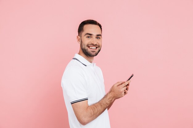 Side view of portrait of a happy handsome man standing isolated over pink , holding mobile phone