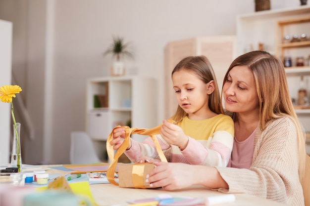 Side view portrait of happy mother hugging daughter while opening presents at wooden kitchen table, copy space