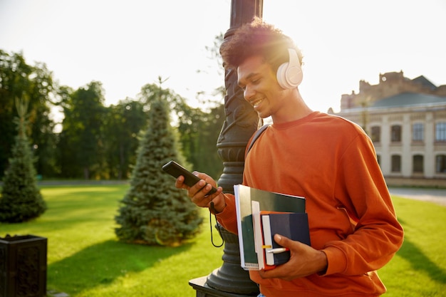 Photo side view portrait of happy smiling teenager student in wireless headset texting on mobile phone walking outdoors at university campus