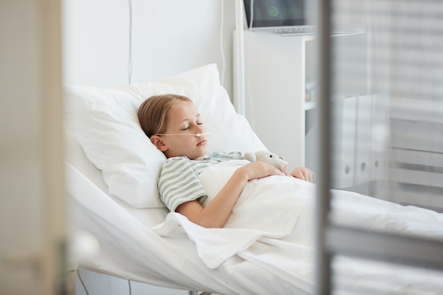 Photo side view portrait of little girl laying in hospital bed with oxygen support, copy space