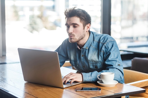 Side view portrait of lovely bearded young handsome man in blue jeans shirt are sitting alone in cafe and making video call sending air kiss to his girlfriend on laptop.