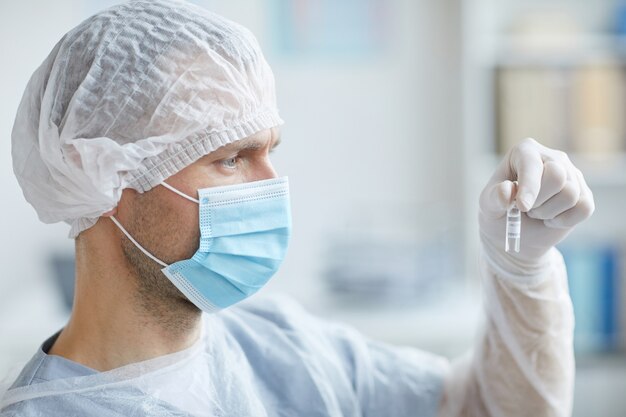 Photo side view portrait of male nurse wearing mask and protective gear holding ampule with medicine while working in clinic or hospital, copy space