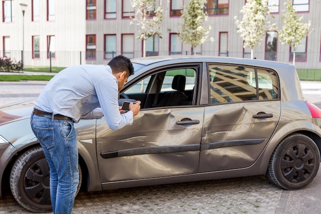 Side view portrait of man wearing jeans and shirt making photo of damaged car after auto accident photographing dents and scratches on vehicle doors and fender Outdoor shot