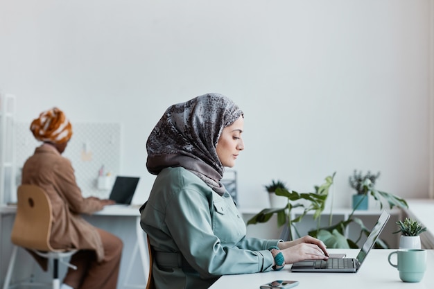 Side view portrait of modern Middle-Eastern woman wearing headscarf in office and using laptop, copy space