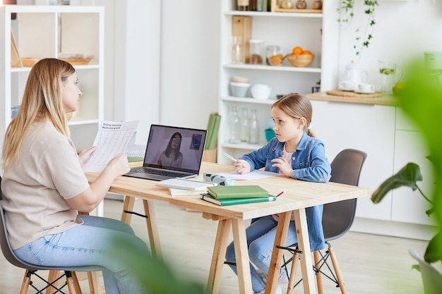 Side view portrait of mother doing homework with cute daughter while sitting at desk and watching online school videos, copy space