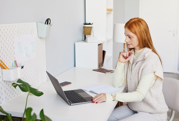 Side view portrait of red haired woman using laptop or studying at minimal home office workplace cop...