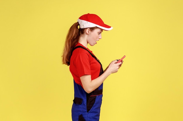 Side view portrait of shocked worker woman using mobile phone, reading message with astonishing news, wearing work uniform and red cap. Indoor studio shot isolated on yellow background.