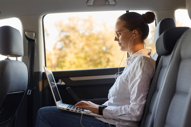 Side view portrait of slim businesswoman wearing white shirt sitting on back seat of car and working with laptop listening music or podcast on her way to office sitting with earphones