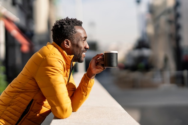 Side view portrait of a smiling African man leaning on a balcony holding a cup of coffee contemplating views in a city