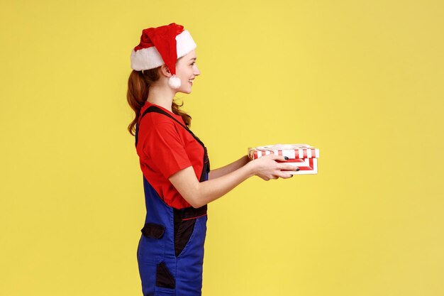 Side view portrait of smiling courier female standing giving wrapped present box for client, wearing blue overalls and santa claus hat. Indoor studio shot isolated on yellow background.