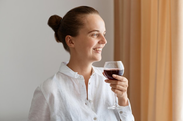 Side view portrait of smiling positive woman wearing white shirt holding glass of red wine looking away with toothy smile expressing happiness celebrating festive event