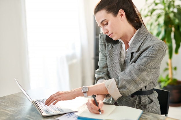 Side view portrait of stressed businesswoman speaking by phone and using laptop while working at desk in office, copy space