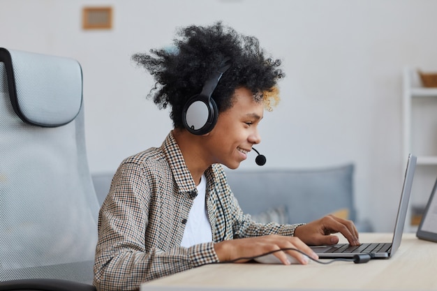 Side view portrait of teenage African-American boy playing video games at home and smiling joyfully, young gamer or blogger concept, copy space