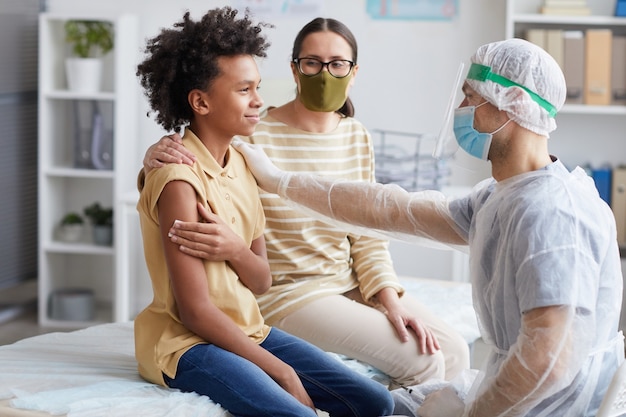 Side view portrait of teenage African-American boy smiling after covid vaccination in clinic with male nurse congratulating him