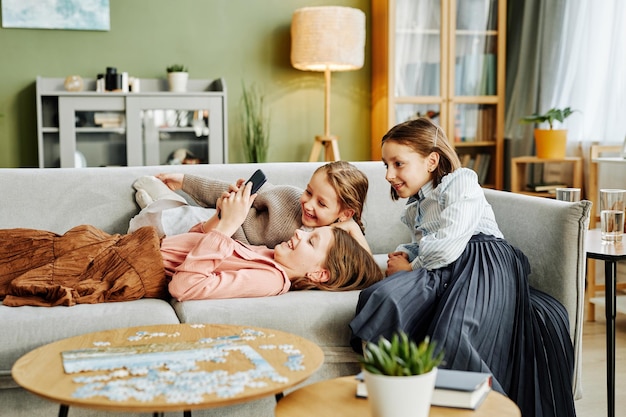 Side view portrait of three little sisters playing together on couch at home and using smart phone