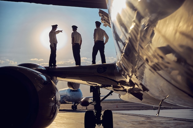 Side view portrait of three unrecognizable persons looking to the sunny sky while standing on the wing of big passenger airplane