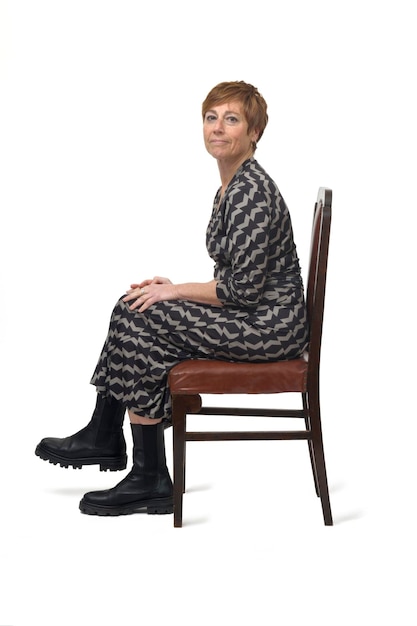 Side view portrait of a woman in dress and boots sitting on chair cross legged and looking at camera on white background