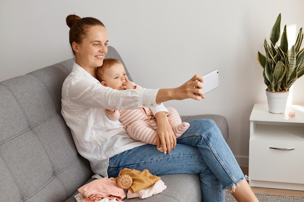 Side view portrait of woman with charming smile sitting on sofa with her toddler baby and holding smart phone in hands, having livestream, talking with her followers.