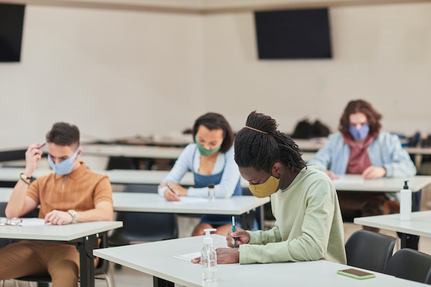 Side view portrait of young African-American man wearing mask while taking test or exam in school with diverse group of people, copy space