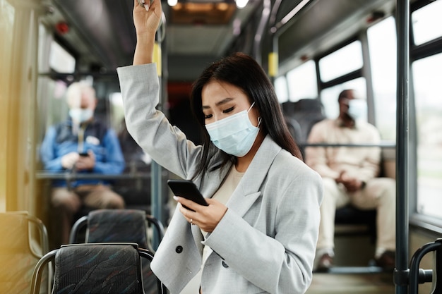 Side view portrait of young Asian woman wearing mask in bus and using smartphone while commuting in city