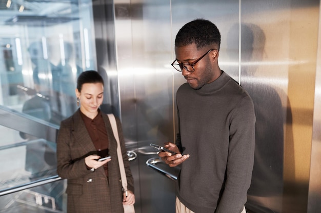 Side view portrait of young black businessman using smartphone in elevator at modern office building