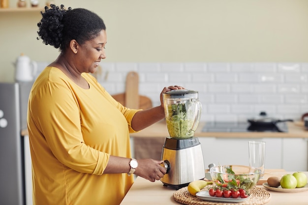 Side view portrait of young black woman using blender while making healthy smoothie at home kitchen