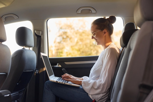 Side view portrait of young business woman using laptop computer while sitting in the back seat of car typing on portable notebook typing important document works online while traveling