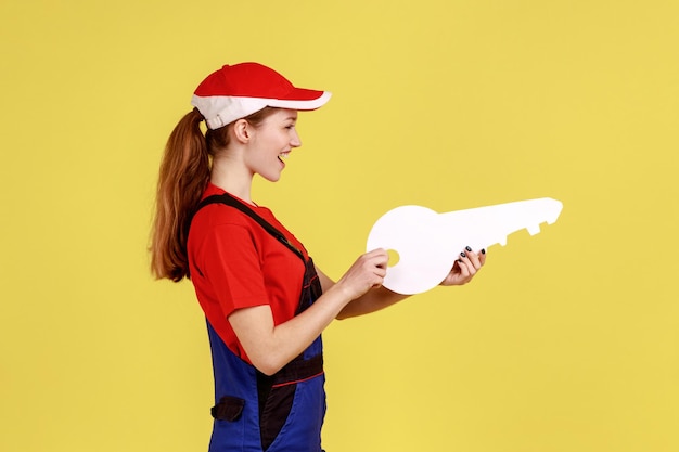 Side view portrait of young happy satisfied worker woman holding big paper key in hands, looking ahead, wearing overalls and red cap. Indoor studio shot isolated on yellow background.