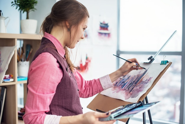 Side view portrait of a young woman painter drawing cityscape with watercolor palette on white paper using easel.