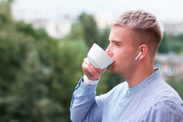 Side view portret of pensive thoughtful blonde guy in shirt, earbuds with cup of coffee or tea. Young man with dyed gray hair listening to music in wireless earphones drinking drink outdoor.