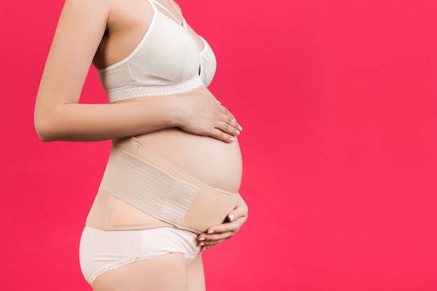 Side view of pregnant woman in underwear wearing pregnancy bandage at pink background with copy space. Close up of orthopedic abdominal support belt concept.