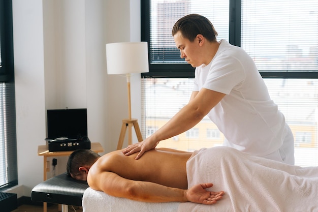 Side view of professional male masseur with strong tattooed hands massaging back and shoulder of muscular sports man lying on stomach at massage table Concept of healthy lifestyle and rest