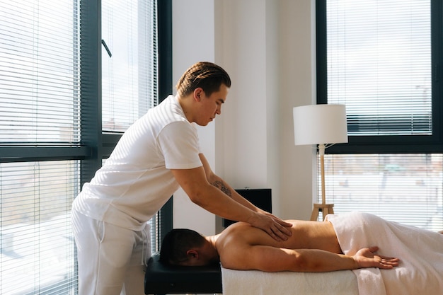 Side view of professional male masseur with strong tattooed hands massaging back and shoulders of muscular sports man lying on stomach