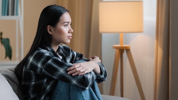 Side view sad asian woman sitting on couch at home feeling unsure about hard decision unhappy