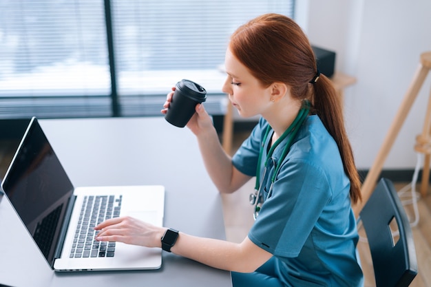 Side view of serious female physician giving distant online consultation to patient via laptop computer and drinking coffee from cup