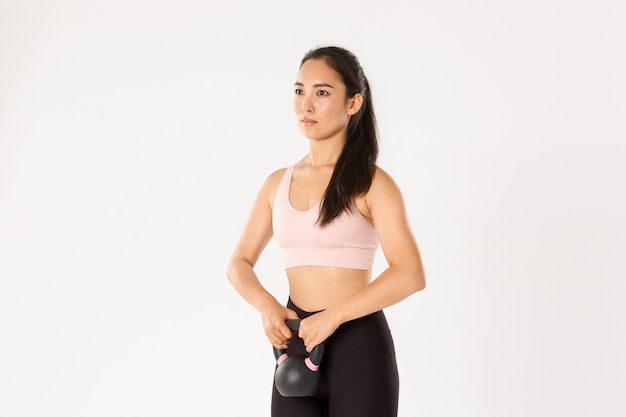 Side view serious and focused asian strong fitness girl, female athlete workout in gym, do squats with kettlebell exercise, standing white background.