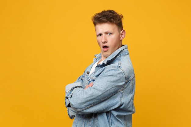 Side view of shocked indignant young man in denim casual clothes holding hands crossed, swearing isolated on yellow orange background. People sincere emotions lifestyle concept. Mock up copy space.