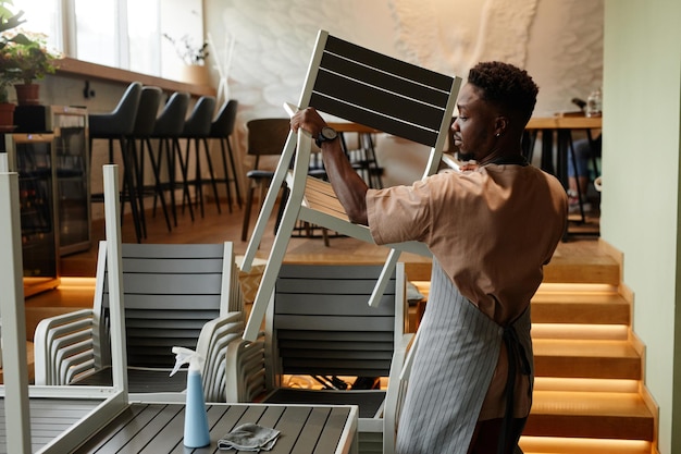 Side view shot of African American man wearing apron starting workday in small cafe preparing tables and chairs for customers before opening