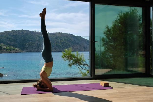 Side view of slim fit female balancing in Supported Headstand pose on mat while doing yoga on terrace in summer