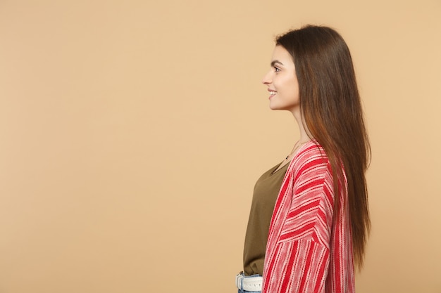 Side view of smiling attractive young woman in casual clothes standing and looking aside isolated on pastel beige background in studio. People sincere emotions, lifestyle concept. Mock up copy space.