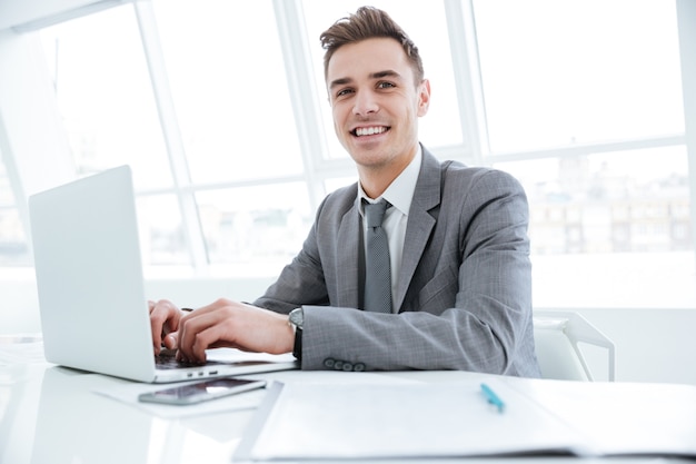 Side view of Smiling business man sitting by the table with laptop computer and looking at camera.